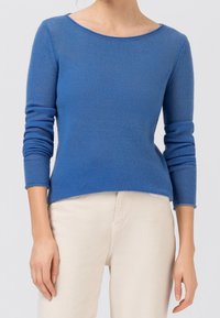 Blauer Strickpullover mit langen Ärmeln und weitem Bootsausschnitt, mit einer gerippten Struktur und einer figurnahen Silhouette. Kombiniert mit hellen Hosen.