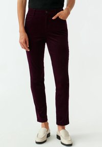 Pantalon en velours côtelé bordeaux avec une coupe ajustée, style cinq poches et fermeture par bouton. Texture lisse, jambe fuselée, porté avec des chaussures beige à enfiler.