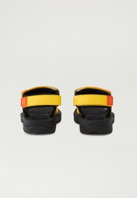 Svarte og gule sandaler med justerbare stropper, som har en teksturert såle og ensfargede detaljer. Bakfra vises en sikker passform-design.