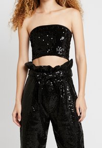 Černý top bez ramínek pokrytý flitry v kombinaci s černými flitrovými kalhotami s vysokým pasem, které mají stahovací pás a volný střih. Hladký a lesklý vzhled po celém outfitu.