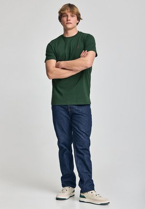 T-shirt verde a maniche corte con scollo rotondo, abbinato a jeans di denim blu scuro e sneakers beige. Vestibilità semplice e casual.