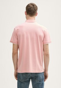 Rosa Polohemd mit kurzen Ärmeln, strukturiertem Stoff und weißem Saum am Kragen und an den Ärmeln. Getragen mit blauen Jeans.