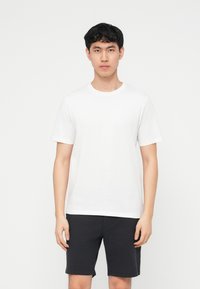 Νεαρός άνδρας που στέκεται μπροστά, φοράει απλό λευκό κοντομάνικο μπλουζάκι και μαύρο σορτς μέχρι το γόνατο, πάνω σε λευκό φόντο.