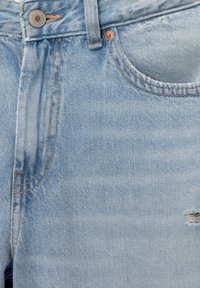 Jean en denim bleu clair en gros plan montrant le bouton, la passants de ceinture, la poche avant, les détails de couture et une petite déchirure du tissu sur le côté droit.