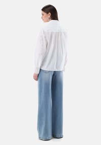 White long-sleeve shirt with eyelet detailing on sleeves and back, paired with light blue wide-leg jeans. Smooth fabric with a relaxed fit.