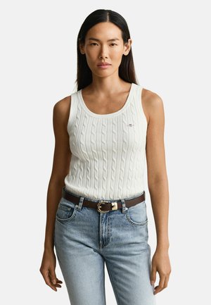 Frau mit langen dunklen Haaren, die ein weißes, ärmelloses, grob gestricktes Oberteil, hellblaue Jeans und einen braunen Gürtel vor einem einfarbigen Hintergrund trägt.