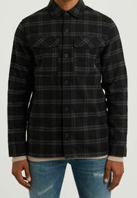Black flannel jacket with gray check pattern, two chest pockets, button closure, and long sleeves. Worn over a light-colored shirt.