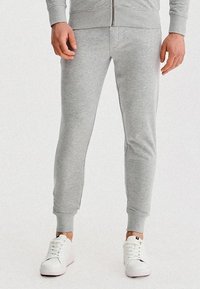 Mężczyzna w jasnoszarych, slim-fit dresach i białych sneakerach, stoj�ący z rękami swobodnie wzdłuż ciała na jednolitym białym tle.