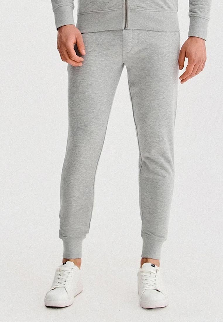 Mężczyzna w jasnoszarych, slim-fit dresach i białych sneakerach, stojący z rękami swobodnie wzdłuż ciała na jednolitym białym tle.