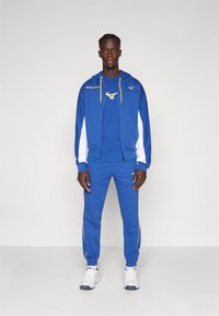 Trening albastru cu un hanorac cu fermoar, având accente albe și galbene, pantaloni asortați cu dungi laterale și pantofi sport albi.