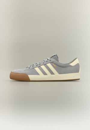 Szary zamszowy sneaker Adidas z kremowymi paskami, gumowym noskiem i podeszwą w kolorze gumy, oznaczony na boku złotym napisem "NORA".