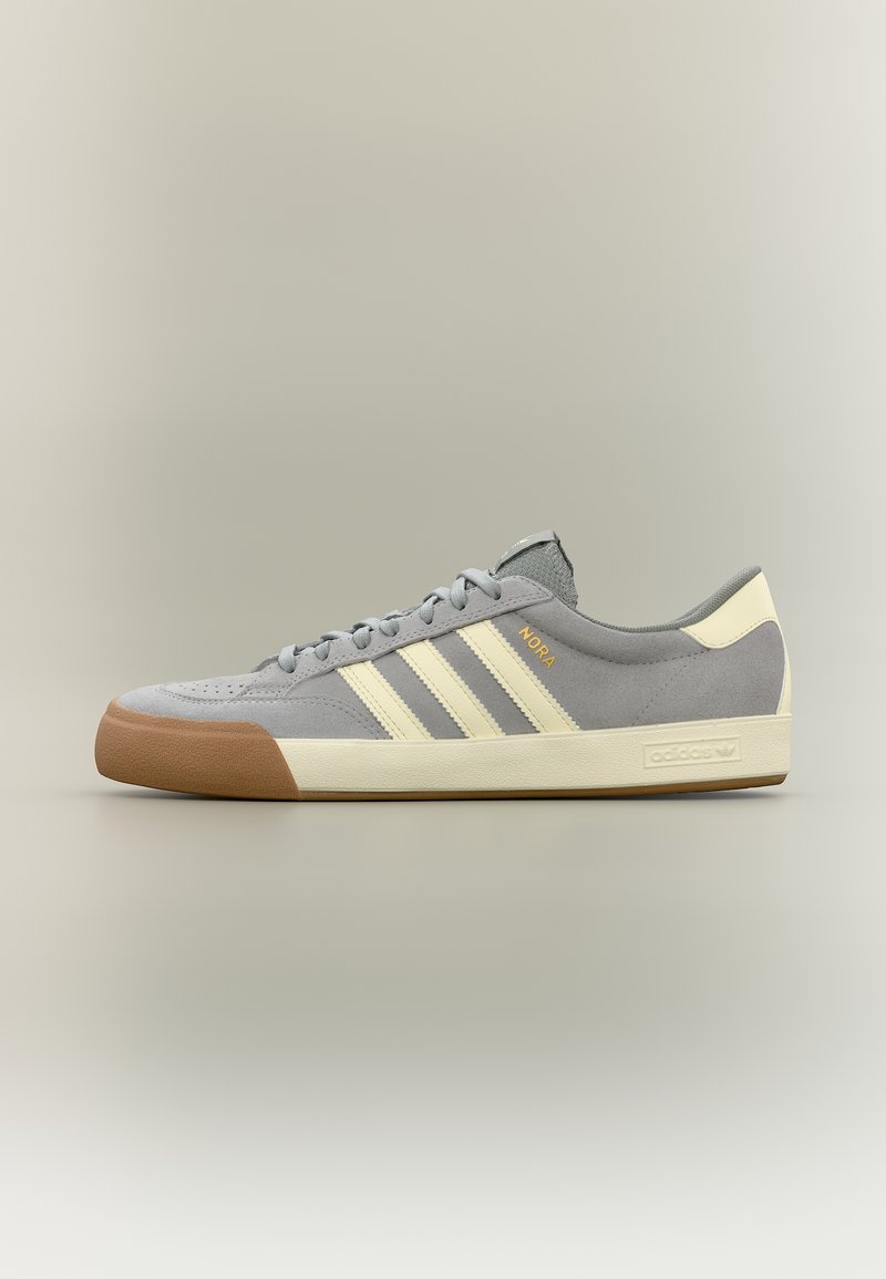 Γκρίζο sneaker Adidas από σουέντ με μπεζ ρίγες, προστατευτικό άκρου δαχτύλων από καουτσούκ χρώματος τσίχλας και σόλα, με την ετικέτα "NORA" σε χρυσό στο πλάι.