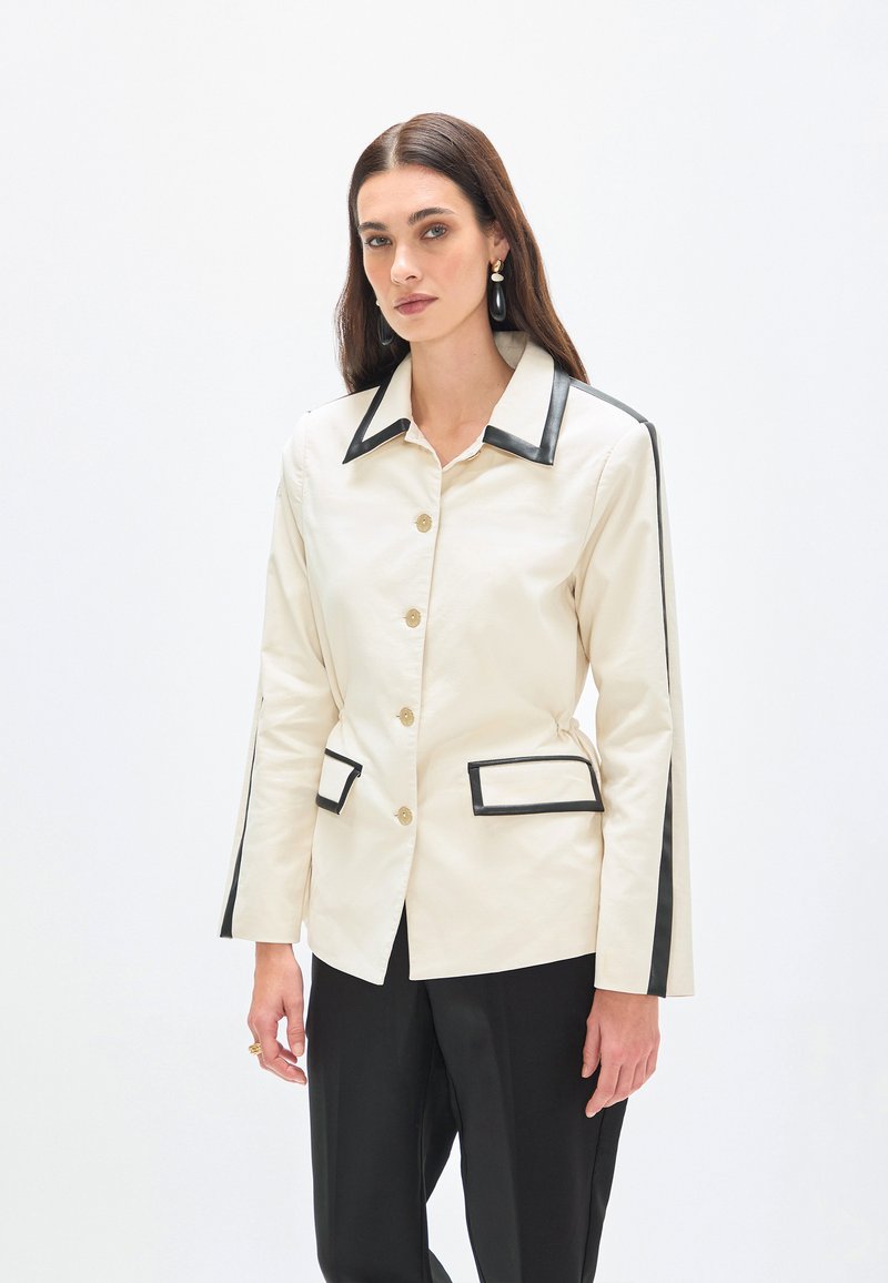 Femme aux longs cheveux foncés portant une veste crème avec un bord noir au col, aux manches et aux poches, associée à un pantalon noir, debout sur un fond blanc.