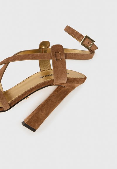 Brūni suede augstpapēžu sandales ar krustveida siksnu dizainu un regulējamu potītes siksniņu, kas ir aprīkota ar zeltītā toņa sprādzi un teksturētu zolīti.