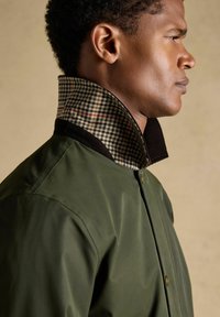 Mann im Profil, der eine olivgrüne Jacke mit hochgestelltem karierter Kragen vor einem schlichten beigefarbenen Hintergrund trägt.