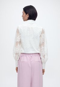 Γυναίκα με κοντά σκουρόχρωμα μαλλιά που φοράει μια λευκή μπλούζα με δαντέλα και λουλουδάτο σχέδιο με φουσκωτά μανίκια και ανοιχτόροζ παντελόνι με ψηλή μέση, γυρισμένη προς τα πίσω.