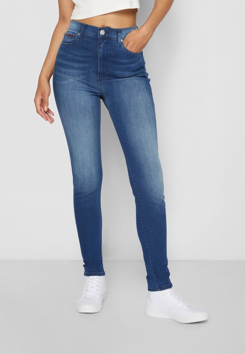 Skinny blue denim jeans with a faded effect, high waist, five-pocket design, and finished with a silver button. Worn with white sneakers.