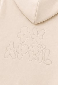 Tissu beige avec texte brodé "Apparil" en grandes lettres bulles ludiques entourées de formes de fleurs simples brodées.