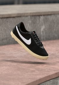 Μαύρο διχτυωτό αθλητικό παπούτσι με λευκό σήμα, επίπεδες κορδόνες και υφή μαύρης μύτης. Διαθέτει κρέμας χρώματος ελαστική σόλα για πρόσφυση.