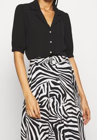 Blouse noire à manches courtes avec col et boutons sur le devant, associée à une jupe blanche arborant un motif audacieux de rayures zébrées noires.