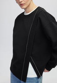 Zwarte sweatshirt met een asymmetrisch ontwerp, voorzien van een ritsdetail langs de zijkant, een zachte textuur en een ronde hals, gelaagd over een wit shirt.