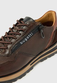Zapato de cuero marrón y textil con una cremallera negra, suela de goma y un logo de la marca. Presenta superficies texturizadas y acentos contrastantes.