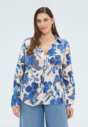 Frau trägt ein blaues Blumenkimono-Hemd mit langen Ärmeln und hellblauen Jeans, steht vor einem einfarbigen hellen Hintergrund.