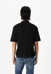 Suéter negro de manga corta con un patrón de canalé texturizado, que presenta un cuello y un diseño ajustado. Lleva puestos unos vaqueros de color azul claro.
