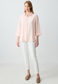 Camicia button-up rosa chiaro con maniche a 3/4, abbinata a pantaloni bianchi slim-fit e scarpe marroni senza lacci. Texture liscia, design minimale.