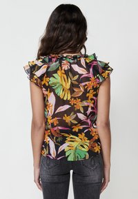 Blusa floreale realizzata in un tessuto nero trasparente, con motivi colorati di foglie e fiori, maniche a volant e scollo arricciato.