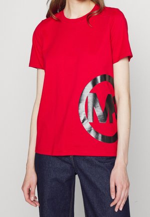 Person wearing a bright red short-sleeve T-shirt with a large black circular logo on the lower left side and dark blue jeans.