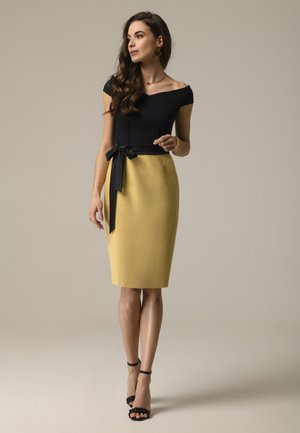 Czarna bluzka bez ramiączek zestawiona z teksturowaną żółtą spódnicą ołówkową, podkreślona czarnym paskiem z materiału i noszona z czarnymi butami na obcasie z odkrytą piętą.