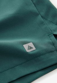 Mørkegrøn atletisk stof med glat tekstur, prydet med et lille gråt logo-tag med et geometrisk design og syede søm-detaljer.