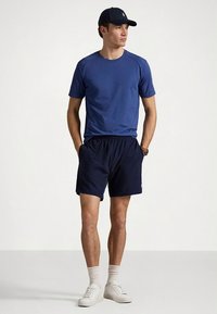 T-shirt atlética azul combinada com calções azuis-escuros, ténis brancos e um boné escuro. A t-shirt tem um ajuste descontraído; os calções apresentam um cós elástico.