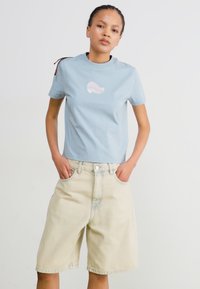 Ljusblå bomulls t-shirt med en rosa grafikdesign, ihop med beiga jeanst-shorts med lös passform och framfickor.