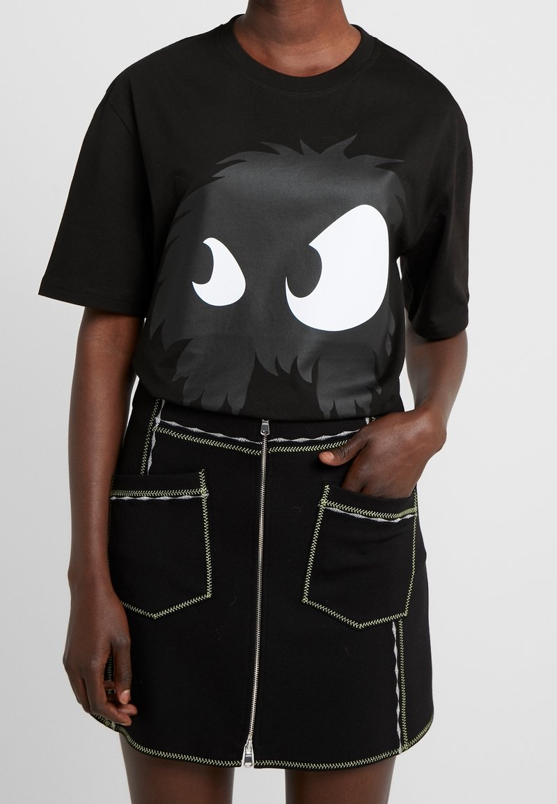 T-shirt in cotone nero con grandi occhi da cartone animato bianchi e un design di capelli testurizzati, abbinato a una gonna nera con cuciture verdi e tasche con zip.