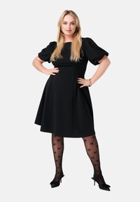 Zwart kort mouwen jurk met pofmouwen, uitlopende rok en knielengte. Gemusterde panty met kleine strikjes; zwarte puntige hakken.