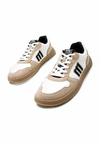 Zapatillas deportivas blancas y beige con textura de gamuza, punta redonda y acentos de logo negro. Presentan un diseño con cordones y suela de goma.