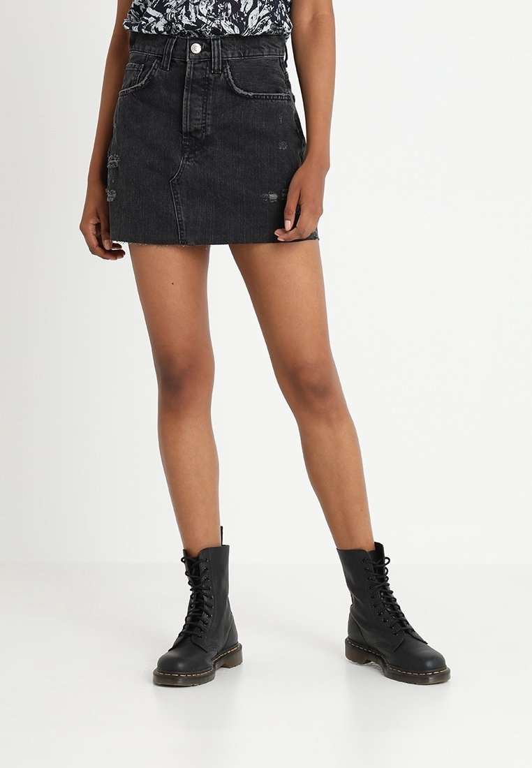 Jupe en denim noir mini, taille haute, avec des détails déchirés et un ourlet brut. Portée avec des bottes de combat noires à lacets.