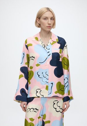 Frau mit kurzen blonden Haaren trägt ein passendes lockeres Hemd und Hose mit großen Blumenmustern in Pink, Blau, Grün und Marinefarben.