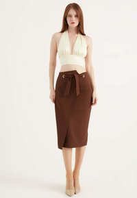 Witte halter crop top met geplooide stof, gecombineerd met een bruine wikkeltule met een strik taille en zilveren oogjes, gedragen met naaktkleurige hakken.