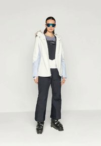 Valge ja helesinine soojustatud jakk halli toppi peal, koos mustade pükste ja mustade suusasaabastega. Mudel kannab tumedaid prille.