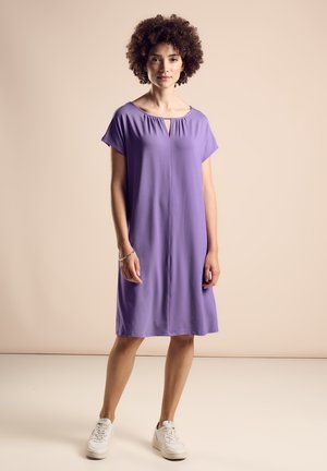 Lyhyet hihat, avainaukolla varustettu violetti mekko, joka on valmistettu pehmeästä kankaasta. Mekko on löysä ja ulottuu polven puoliväliin, yhdistettynä valkoisiin tennareihin.