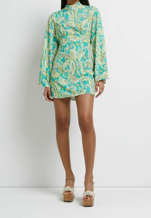 Mujer con un vestido corto turquesa con estampado paisley, mangas largas y sandalias de plataforma tostadas con tiras trenzadas blancas.