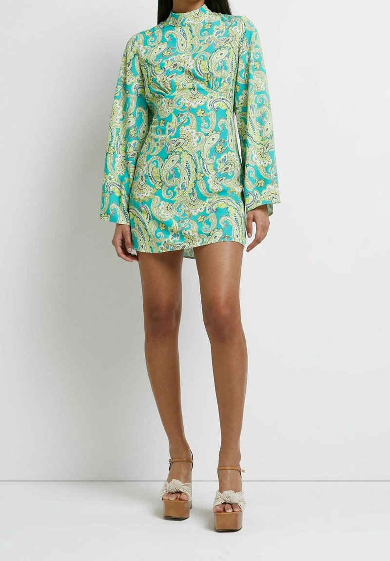 Femme portant une courte robe turquoise à motif cachemire, manches longues, et des sandales compensées couleur cuir clair avec des brides tressées blanches.