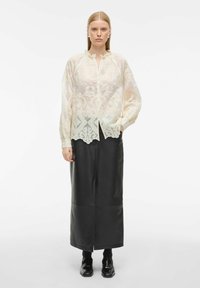 Gennemsigtig, cremet bluse med dekorativ broderi i kanten, parret med en tætsiddende sort læderskirt, og sorte fodtøj med en blank finish.