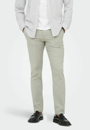 Man die lichtgrijze slim-fit broek, wit T-shirt, lichtgrijs gestreept overhemd met knopen en zwarte leren loafers draagt, met zijn handen in zijn zakken.