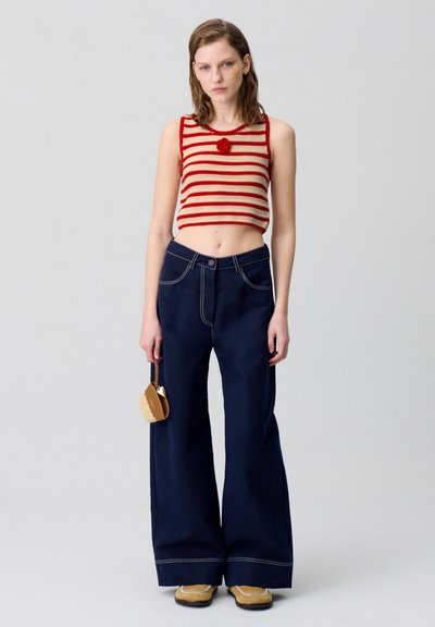 Top corto a righe rosso e crema con dettagli floreali, abbinato a pantaloni larghi blu scuro e scarpe marroni, mentre tiene una piccola borsa marrone.