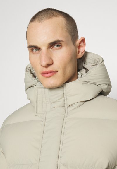 Beige pufferjakke med høj krave og hætte, prydet med en quiltet design og lynlås-lukning. Glat tekstur med minimal hardware.