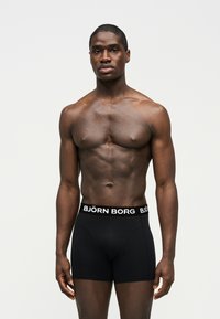 Homme musclé debout, portant un boxer noir Björn Borg, sur un fond clair uni, faisant face à l'avant avec les bras détendus.